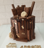 Tall chocolate drip cake loaded with chocolate bars, gold macarons, and edible gold leaf on a white marble surface.
