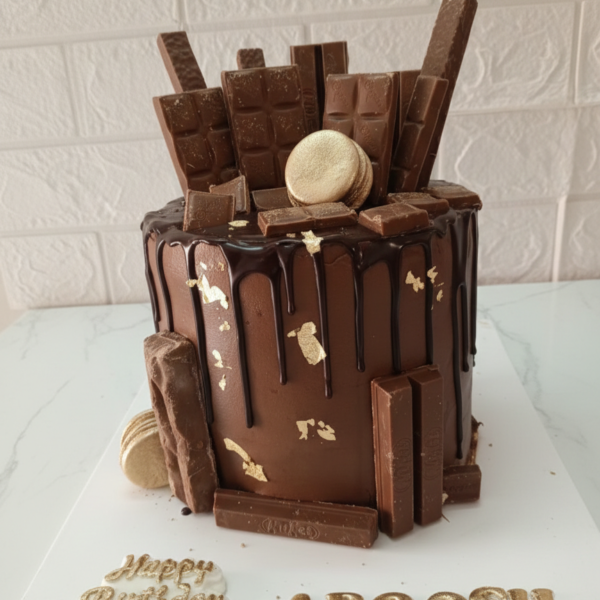 Tall chocolate drip cake loaded with chocolate bars, gold macarons, and edible gold leaf on a white marble surface.