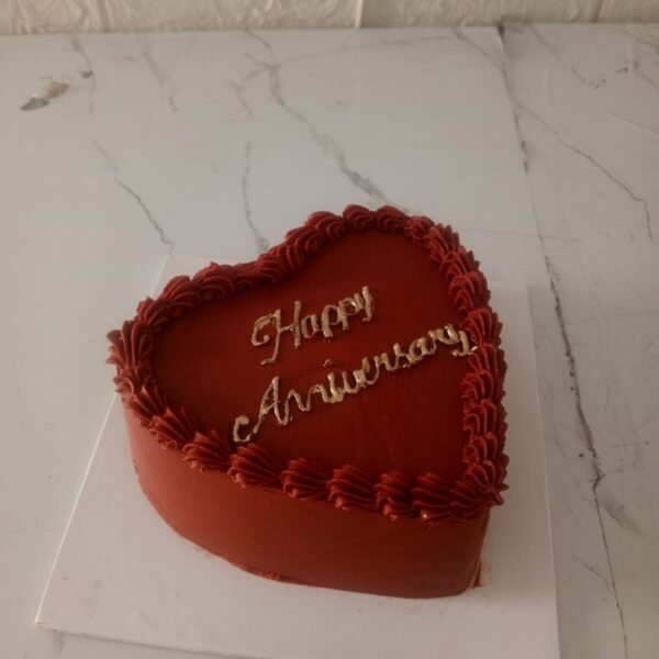 Heart-shaped red anniversary cake with golden "Happy Anniversary" topper on a white marble background.