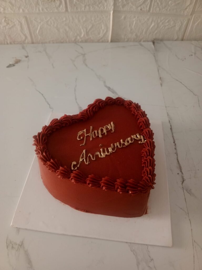 Heart-shaped red anniversary cake with golden "Happy Anniversary" topper on a white marble background.