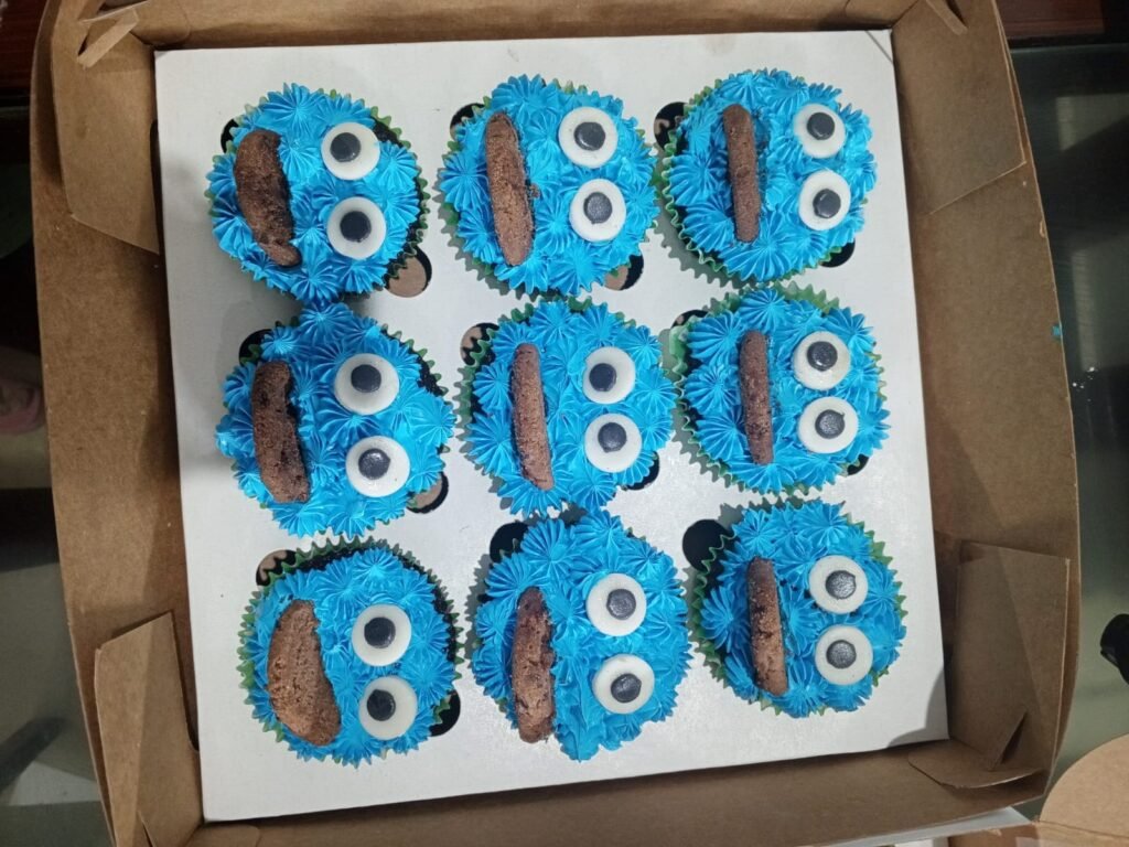 A box of nine blue Cookie Monster cupcakes with googly eyes and chocolate chip cookies.